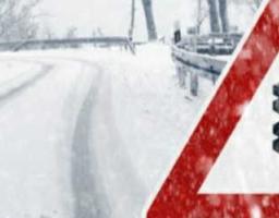 На зображенні великим планом показано червоний трикутний попереджувальний дорожній знак із чорною сніжинкою всередині. Цей знак, що належить до попереджувальних знаків, інформує водіїв про небезпеку попереду, зокрема про слизьку дорогу через сніг або ожеледицю. На задньому плані видно засніжену, слизьку ділянку дороги та засніжені дерева