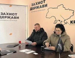 На зображенні двоє людей, чоловік і жінка в зимовому одязі, сидять за столом у кімнаті. Вони тримають папери, а на столі перед ними лежить інша документація, ручка, печатка та невеличкий чорний прапорець з написом «ЗАХИСТ ДЕРЖАВИ». На стіні позаду них розташовані банери з великими написами «ЗАХИСТ ДЕРЖАВИ» та контуром карти України
