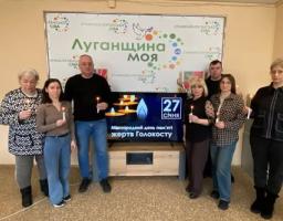 На зображенні показано групу з шести людей (три жінки та троє чоловіків), які стоять у приміщенні та тримають у руках запалені свічки. Вони стоять перед великим банером із написом «Луганщина моя». Під банером розташований телевізор, на екрані якого відображається напис «Міжнародний день пам'яті жертв Голокосту» та дата «27 СІЧНЯ» разом із зображенням свічок