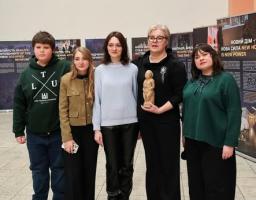 Зображення показує п'ятьох людей, які стоять поруч у приміщенні галереї чи музею з інформаційними банерами на задньому плані, одна з жінок тримає невелику статуетку.