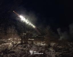 Зображення показує нічну сцену з військовою технікою (пусковою установкою), яка веде вогонь. На темному тлі видно яскравий спалах пострілу та димовий слід від ракети, що летить угору вліво. Земля вкрита снігом та сухою рослинністю