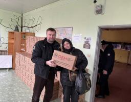Зображення є фотографією двох людей, ймовірно, волонтерів, які тримають картонну коробку з гуманітарною допомогою. На коробці видно логотип «GEM» (Global Empowerment Mission) та напис "UKRAINE", а також адресу веб-сайту globalempowermentmission.org