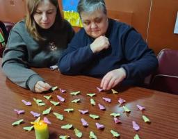 На зображенні дві жінки сидять за дерев'яним столом і працюють із численними маленькими паперовими фігурками (схожими на метеликів або сердечка) рожевого та світло-зеленого кольорів. Фігурки розкидані по поверхні столу. На передньому плані ліворуч стоїть невелика запалена свічка у зеленій підставці.