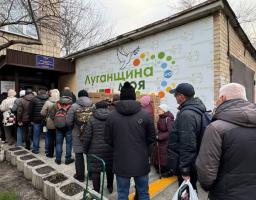 На зображенні видно чергу людей, одягнених у зимовий одяг, які стоять перед будівлею з великим банером із написом «Луганщина моя», ймовірно, очікуючи на отримання гуманітарної допомоги або послуг.