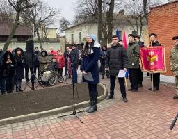 На зображенні видно натовп людей, які зібралися на офіційній церемонії на відкритому повітрі, де жінка виступає біля мікрофона, а молоді люди у формі тримають прапори.