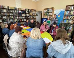 Група з приблизно десяти людей, переважно жінок, сидить навколо яскравого столу в бібліотеці, де на задньому плані видно книжкові полиці та предмети, схожі на військову техніку, слухаючи жінку, яка стоїть біля дошки
