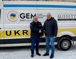 На зображенні двоє чоловіків тиснуть руки перед білим фургоном із логотипами GEM Ukraine Global Empowerment Mission та The Howard G. Buffett Foundation на сніжному тлі.
