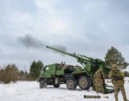 Зображення показує велику зелену самохідну гаубицю CAESAR на колесах, яка стріляє в засніженому лісовому ландшафті, оточена кількома солдатами у військовій формі.