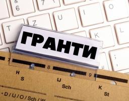 На зображенні показано архівну теку або папку-реєстратор із роздільниками, на якій розміщена настільна табличка зі змінним написом великими чорними літерами: "ГРАНТИ". Фон зображення становить світла клавіатура.