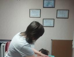 Зображення показує, як фахівець проводить сеанс масажу спини клієнту на масажному столі, при цьому на стіні позаду висять декілька сертифікатів або дипломів у рамках