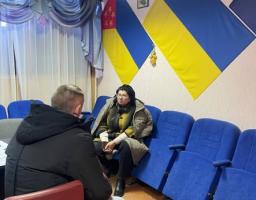 Зображення показує двох людей, що сидять обличчям один до одного під час зустрічі в офіційній кімнаті, прикрашеній прапорами України та регіональним прапором, а також рядами синіх крісел.