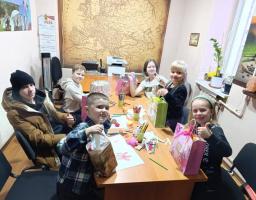 Шестеро дітей різних вікових груп радісно сидять за столом в офісі, де на стіні висить велика карта, тримають подарункові пакети та демонструють великі пальці вгору.