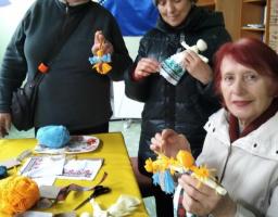 На фото зображено трьох жінок, які під час волонтерського майстер-класу в Закарпатті власноруч створюють традиційні українські ляльки-мотанки та патріотичні обереги з жовто-блакитної пряжі та вишитого полотна, символізуючи єдність та незламність національного духу