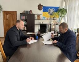 Двоє чоловіків підписують документи за столом в офісі, де на стіні висить український прапор.