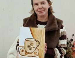 На зображенні усміхнена жінка тримає свій малюнок, виконаний у теплих коричневих тонах. На малюнку зображена чашка з квітковим візерунком на блюдці, що стоїть на столі біля завіси. Жінка одягнена в хутряну жилетку поверх смугастого джемпера, а на голові в неї сонцезахисні окуляри