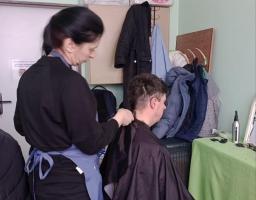 Жінка в чорній кофті та блакитному фартуху стриже чоловіка в чорному перукарському пеньюарі в невеликому приміщенні, де на задньому плані видно складені коробки, підвішений верхній одяг та перукарське приладдя