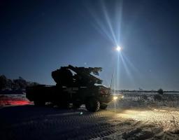 Військова вантажівка, оснащена зенітно-ракетним комплексом, стоїть у засніженому полі в нічний час, освітлена яскравим місяцем та фарами