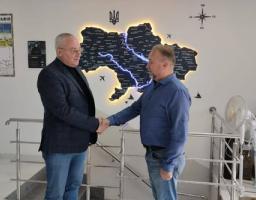 На зображенні представлено двоє чоловіків у приміщенні, які тиснуть один одному руки на фоні стіни з картою України та плакатом з військової топографії. Чоловік ліворуч одягнений у темно-синій піджак, чорний гольф та джинси, а чоловік праворуч — у синю сорочку та джинси. Зустріч відбувається біля сходової клітки з металевими поручнями, а поруч на підлозі стоять предмети, схожі на тубуси для зброї або обладнання