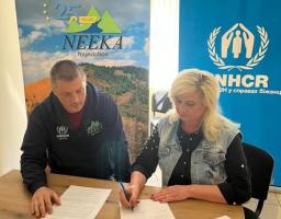 Чоловік та жінка в одязі з корпоративною символікою зосереджено підписують офіційні документи, сидячи за дерев'яним столом.