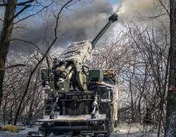 Українська самохідна артилерійська установка 2С22 «Богдана» веде вогонь по позиціях противника в зимовому лісі. Ствол гармати піднятий вгору, з нього виривається дим після пострілу; установка частково вкрита білою маскувальною сіткою.