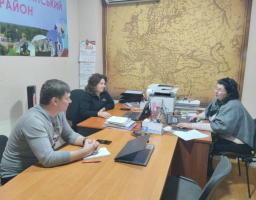 Три людини, двоє чоловіків та жінка, сидять за дерев'яним столом в офісі, де на стіні висить велика карта Європи та плакат з написами