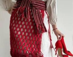 Жінка в етностилі: біла вишита сорочка та довга спідниця, доповнені широким тканим поясом і червоною макраме-запаскою (припиндою) з бахромою. В одній руці вона тримає пару класичних червоних туфель на шпильці.