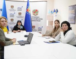 Чотири жінки проводять офіційну зустріч або брифінг у приміщенні «Official Media Center» Луганської обласної військової адміністрації. Вони сидять за великим білим столом, на якому стоять ноутбук та документи. На задньому плані розміщено прапор Луганської області, логотипи медіацентру та інформаційний плакат для ветеранів