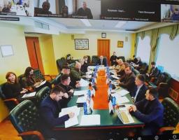 На екрані ноутбука відображено робочу зустріч: група посадовців сидить за великим столом у залі засідань, а у верхній частині вікна видно іконки учасників відеоконференції. На столі лежать папери, ноутбуки та пляшки з водою