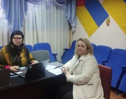Дві жінки проводять робочу зустріч в офісному приміщенні. Одна сидить за столом із ноутбуком, інша — поруч із документами. На стіні позаду розміщені прапори України та Луганської області