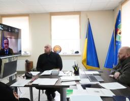 За столом сидить двоє чоловіків у темному одязі та худі кольору хакі, а також жінка (вид зі спини). На великому настінному екрані відображається відеодзвінок з іншим учасником