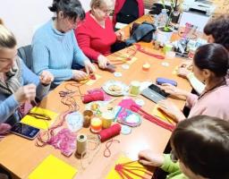 Група жінок за довгим столом займається рукоділлям, імовірно, плетінням макраме з червоних ниток. На столі розкладені заготовки, нитки, інструменти та частування; на стіні висить календар на 2026 рік.
