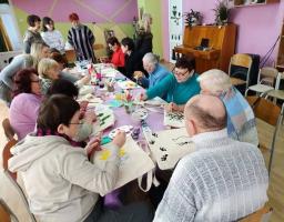 Група людей різного віку, переважно похилого, зібралася в яскравій кімнаті для творчого майстер-класу. Вони сидять за довгим столом і розмальовують фарбами екосумки. На фоні видно піаніно, картини на стінах та інших учасників, які спостерігають за процесом.