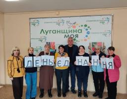 Група людей стоїть у приміщенні на фоні банера з написом «Луганщина моя» та логотипами Станично-Луганської СВА. У руках вони тримають аркуші паперу, які разом утворюють напис «ДЕНЬ ЄДНАННЯ».