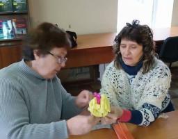 Дві жінки старшого віку, які зосереджено працюють над створенням виробу. Одна з них показує іншій, як правильно формувати деталь