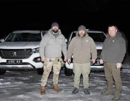 На зображенні — троє чоловіків у тактичному та повсякденному одязі, які стоять на снігу на фоні двох білих пікапів