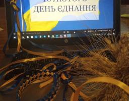 На ноутбуці відкрите зображення з написом «16 ЛЮТОГО ДЕНЬ ЄДНАННЯ» на фоні українського прапора. Перед клавіатурою лежать синьо-жовті стрічки, плетений браслет і пучок колосків пшениці.