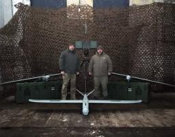 На фото зображено двох чоловіків, які стоять поруч із безпілотними авіаційними комплексами (БпАК)