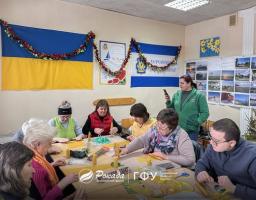 Група людей на творчому майстер-класі від Благодійного фонду «Рокада». Учасники сидять за столом у світлій кімнаті, прикрашеній українськими прапорами, та виготовляють вироби ручної роботи