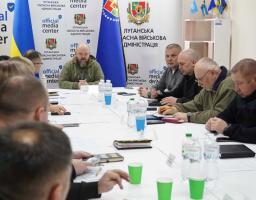 За довгим білим столом зібралася група людей. У центрі (на фоні прапорів) — чоловік у військовій формі оливкового кольору, який, ймовірно, головує на зустрічі. Праворуч від нього сидять представники різних відомств, зокрема рятувальник ДСНС (у чорній формі з нашивкою)