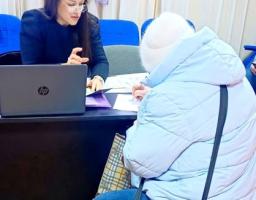 За столом сидить жінка-спеціаліст у діловому одязі та окулярах, вона щось пояснює та вказує на документи. Навпроти неї, спиною до камери, сидить відвідувачка у світлому зимовому одязі (пуховику та шапці), яка в цей момент підписує папери