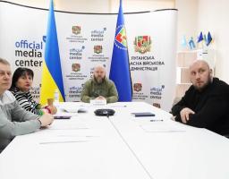 Представники Луганської обласної військової адміністрації проводять брифінг в офіційному медіацентрі на фоні державних прапорів та інформаційних стендів