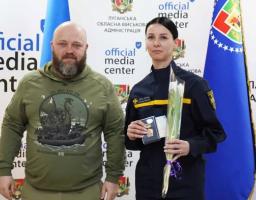 Чоловік у худі кольору хакі та жінка у формі ДСНС із квітами й нагородою позують на фоні банера Луганської обласної військової адміністрації