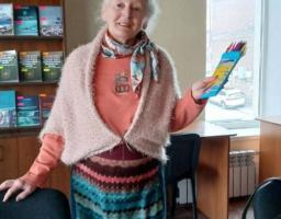 Усміхнена жінка похилого віку в колоритному в'язаному вбранні стоїть у бібліотеці чи класі, тримаючи в руках набір кольорових олівців