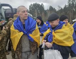На фото зображено звільнених з полону українських захисників, які стоять на вулиці, загорнуті в національні прапори України