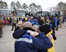 Група чоловіків, дехто з яких загорнутий в українські прапори, емоційно обіймаються на тлі автобусів під час обміну полоненими
