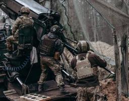 Троє українських артилеристів у повному спорядженні працюють біля розгорнутої артилерійської установки під маскувальною сіткою в лісистій місцевості.