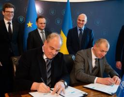На офіційному заході двоє чоловіків у ділових костюмах підписують документи за столом, у той час як за ними стоять інші офіційні особи на фоні прапорів України та Європейського Союзу
