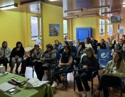 Група людей уважно слухає доповідача під час інформаційної зустрічі або тренінгу в жовтій аудиторії, де на столі перед ними розкладені блокноти та матеріали для записів.