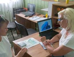 Дві жінки в білих футболках сидять за робочими столами одна навпроти одної в офісному приміщенні зі світло-зеленими стінами та ноутбуками, обговорюючи документи під час робочого процесу.
