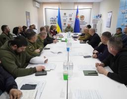 На світлині зображено офіційне засідання групи посадовців та військових, які сидять за довгим білим столом у конференц-залі «Official Media Center» на фоні державних прапорів України